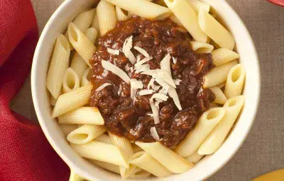 Fotografia em tons de amarelo e vermelho, ao centro uma panela destampada com o macarrão tipo penne e o ragú em cima decorado com queijo ralado. Ao lado está a tampa amarela da panela e um pote vermelho com queijo ralado.